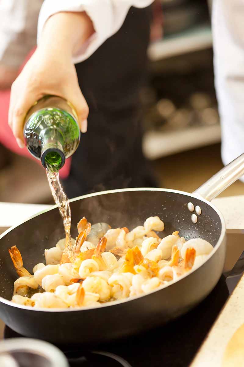 Vertical image of pouring a liquid from a bottle into a pan with shrimp on the stovetop.