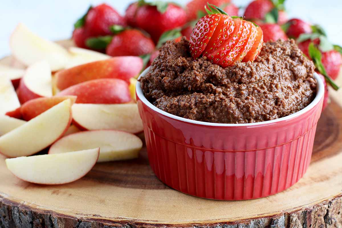 Horizontal image of a chocolate and chickpea recipe in a red ramekin on a wooden platter next to apple wedges and whole strawberries.