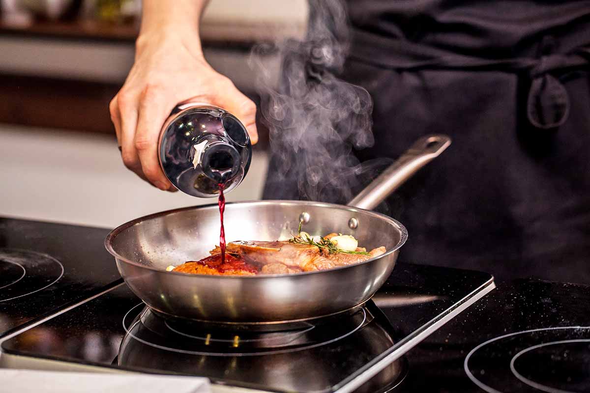 Horizontal image of pouring red-colored alcohol from a bottle into a skillet with meat searing on the stovetop.
