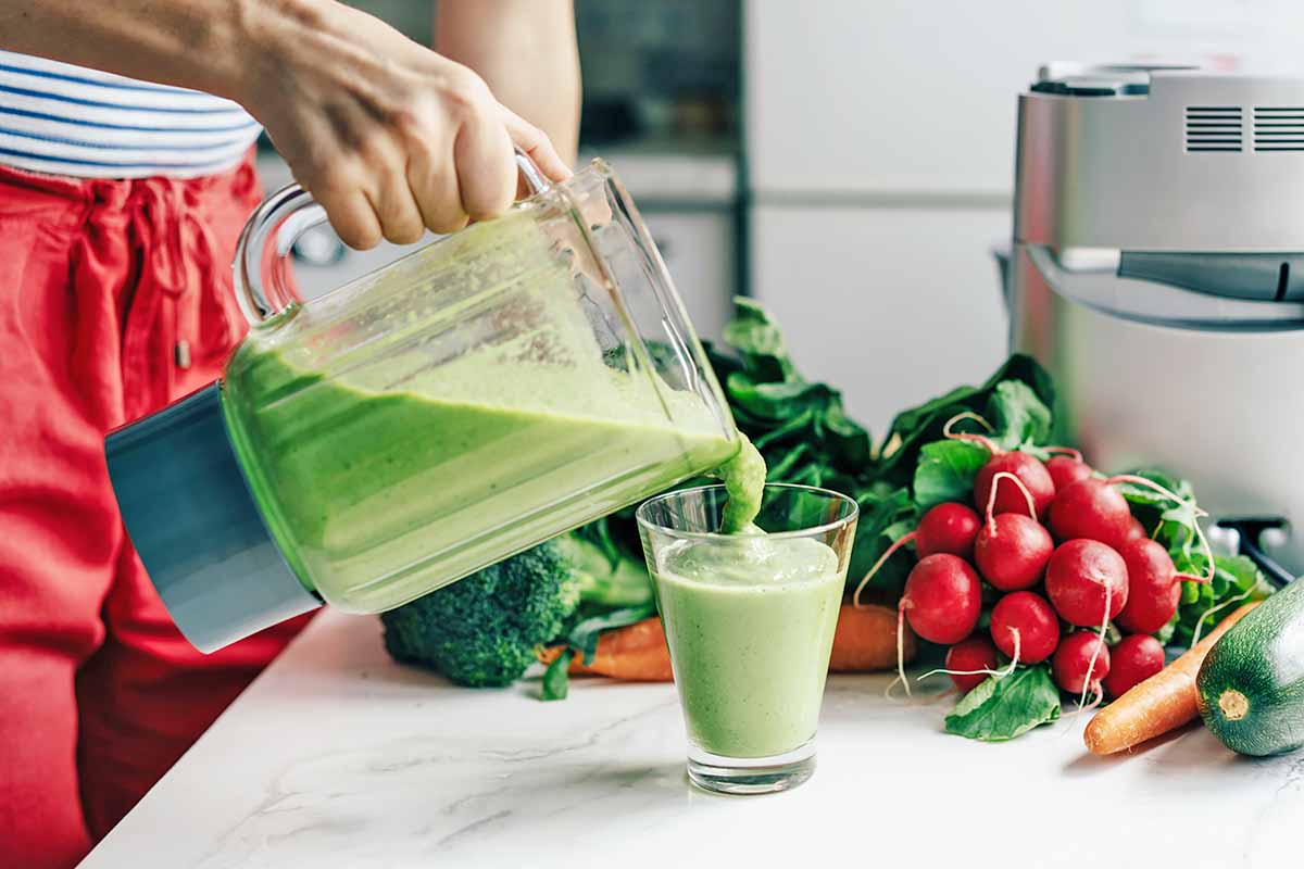 Horizontal image of a woman pouring a green creamy shake into a glass cup on a countertop with assorted vegetables.