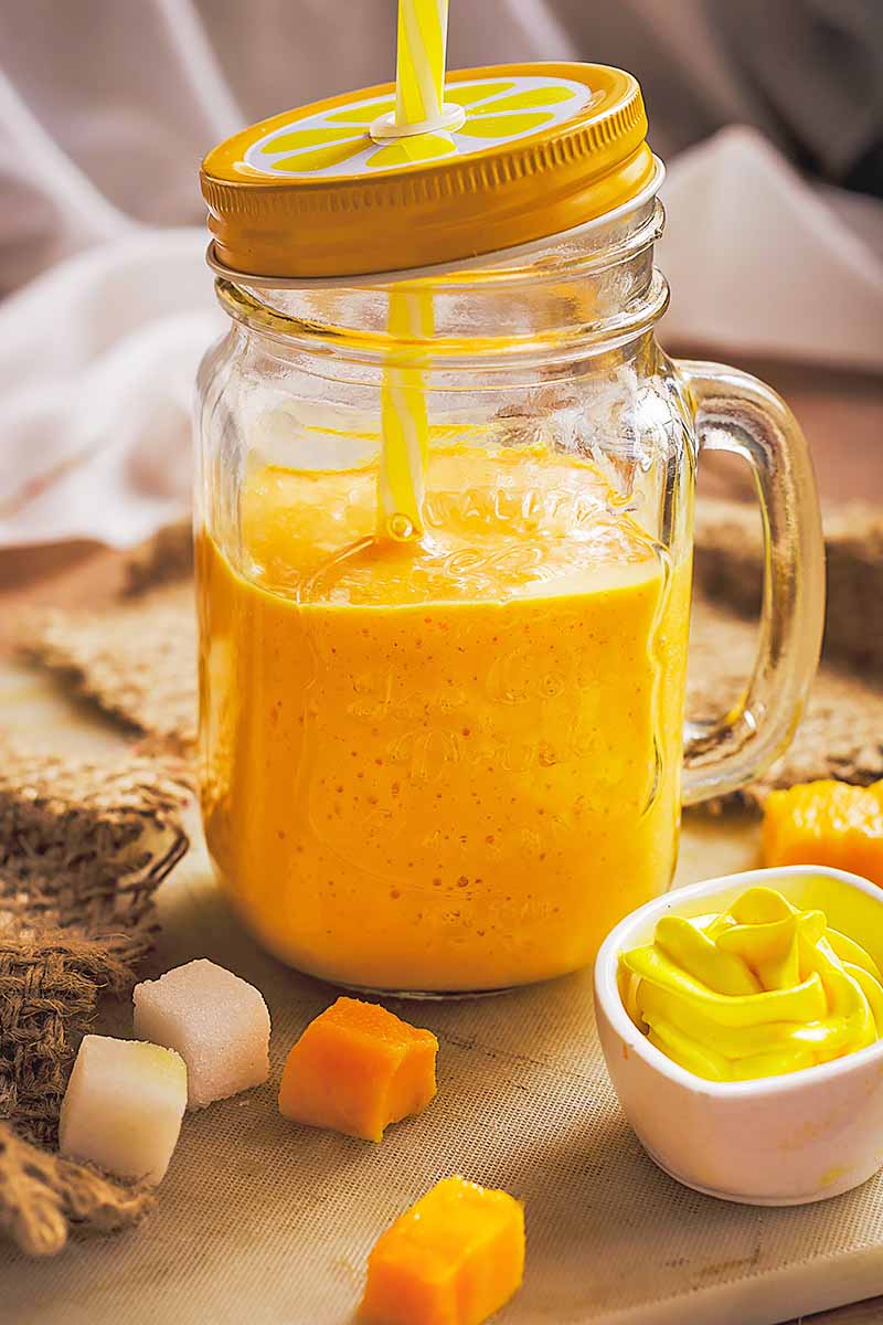 Vertical image of an orange shake in a glass mason jar with a lid and straw on a counter with assorted ingredients and a burlap napkin.
