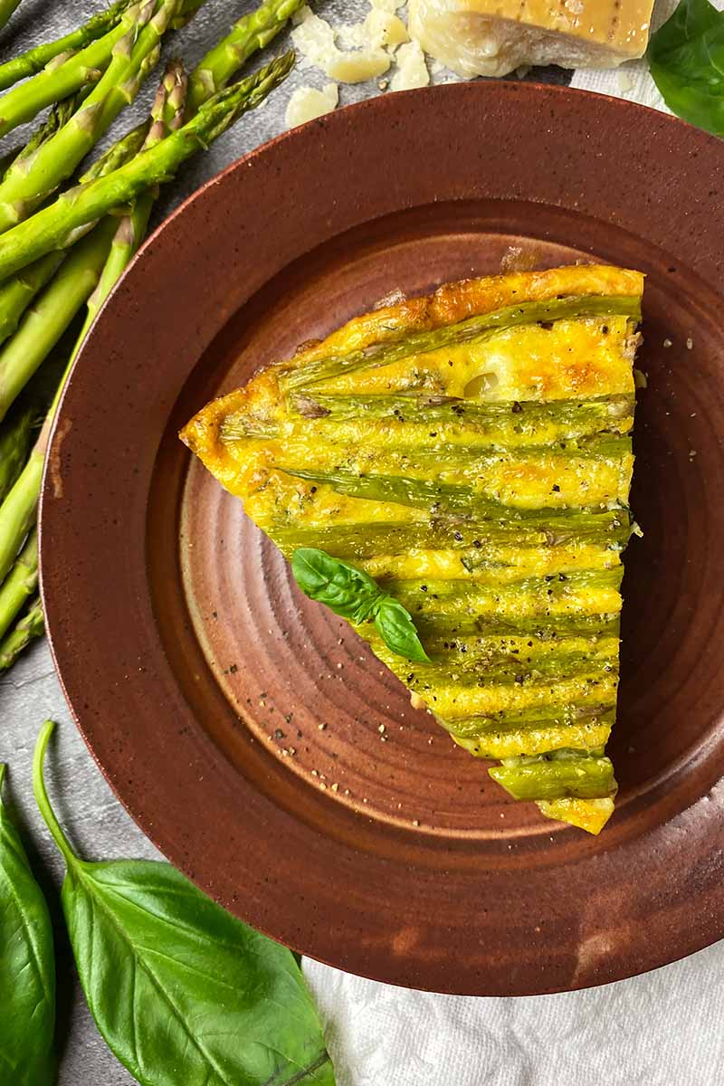 Vertical top-down image of a wedge of an asparagus frittata garnished with basil on a brown plate.