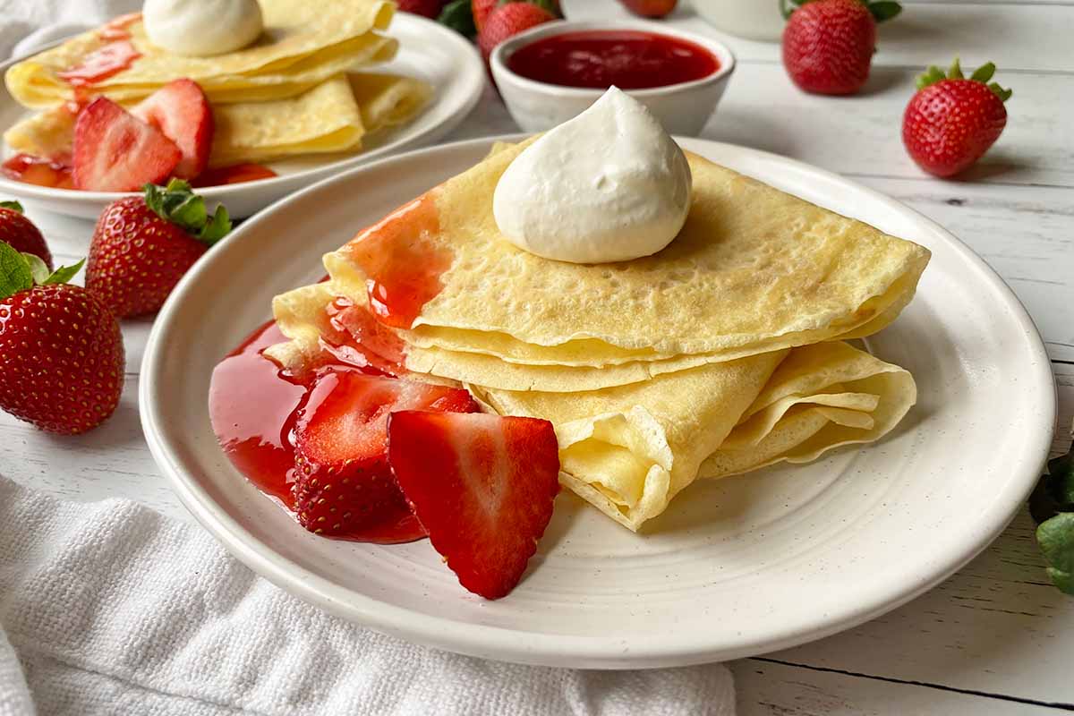 Horizontal image of two white plates, each with with a stack of folded thin pancakes served with whipped cream, sliced fruit, and a red syrup.