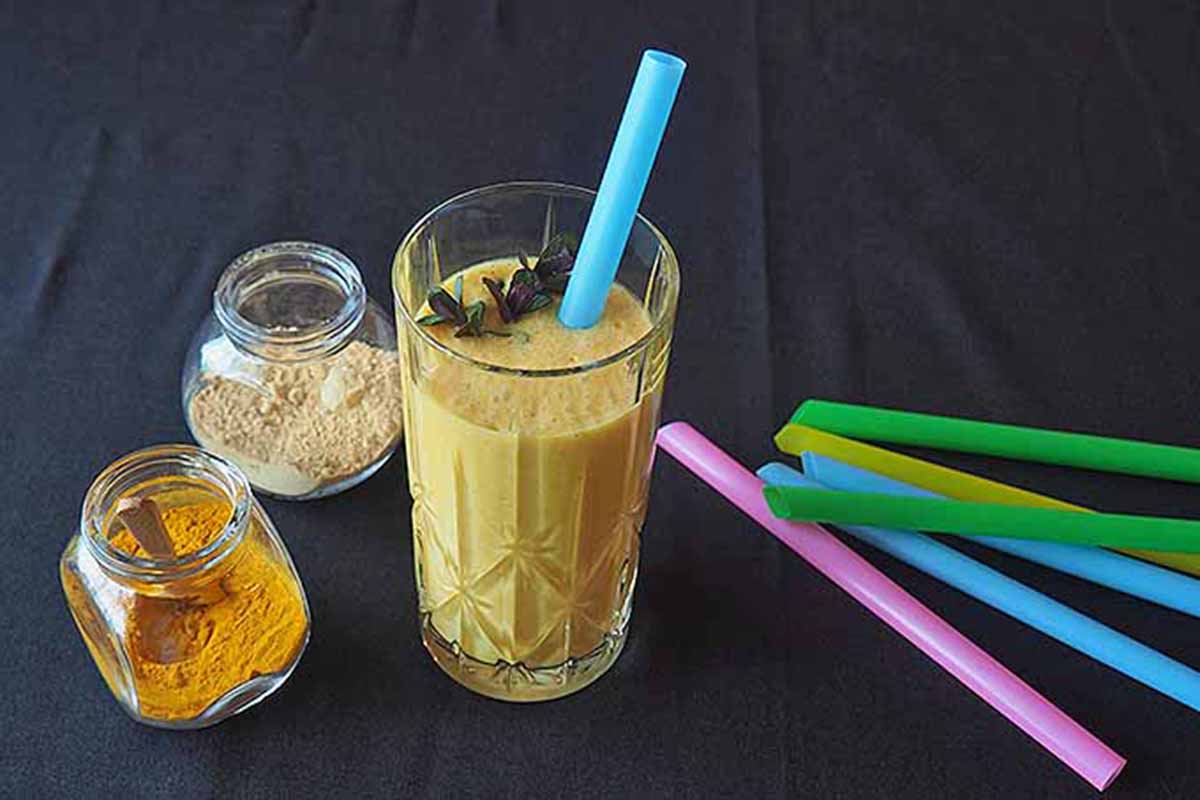 Horizontal image of a creamy pineapple drink next to glasses of spices and straws on a black tablecloth.