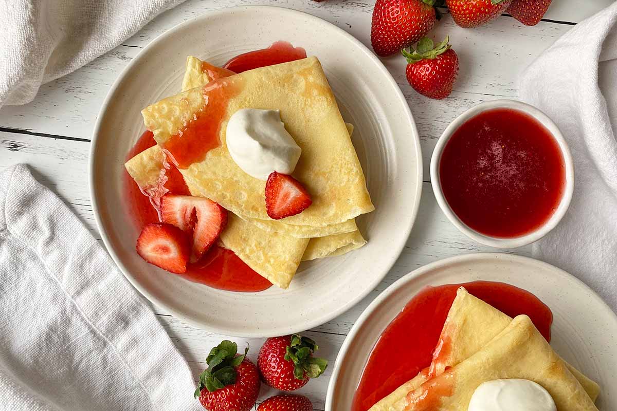 Horizontal top-down image of two white plates, each with with a stack of folded thin pancakes served with whipped cream, sliced fruit, and a red syrup.
