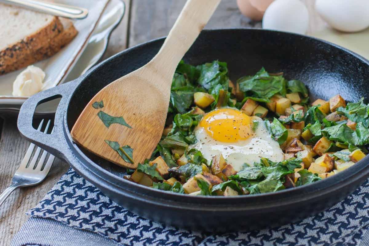 Horizontal image of a beet green and potato hash in a skillet with a wooden spoon on a towel.