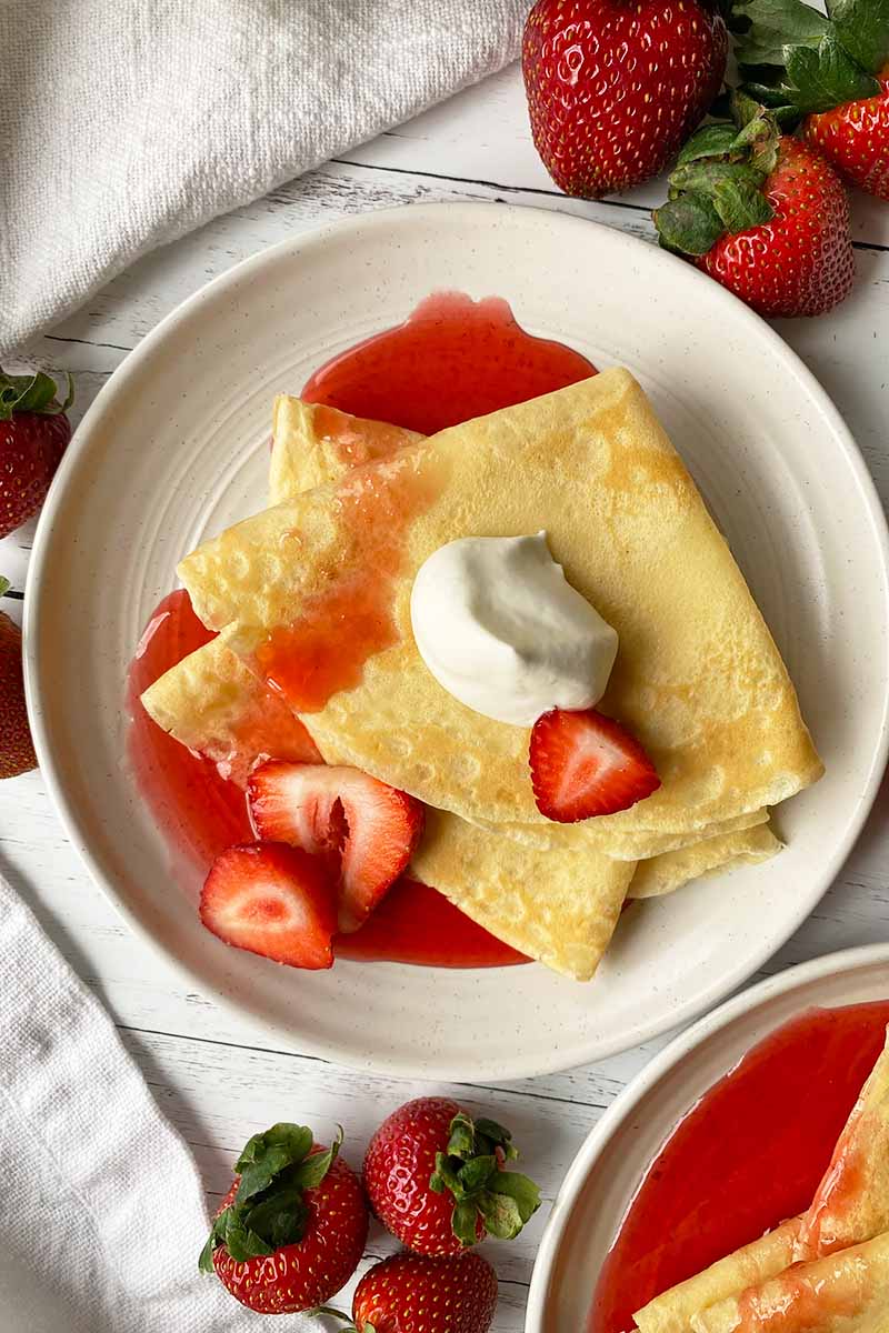 Vertical top-down image of a white plate with a stack of folded thin pancakes served with whipped cream, sliced strawberries, and a red syrup.