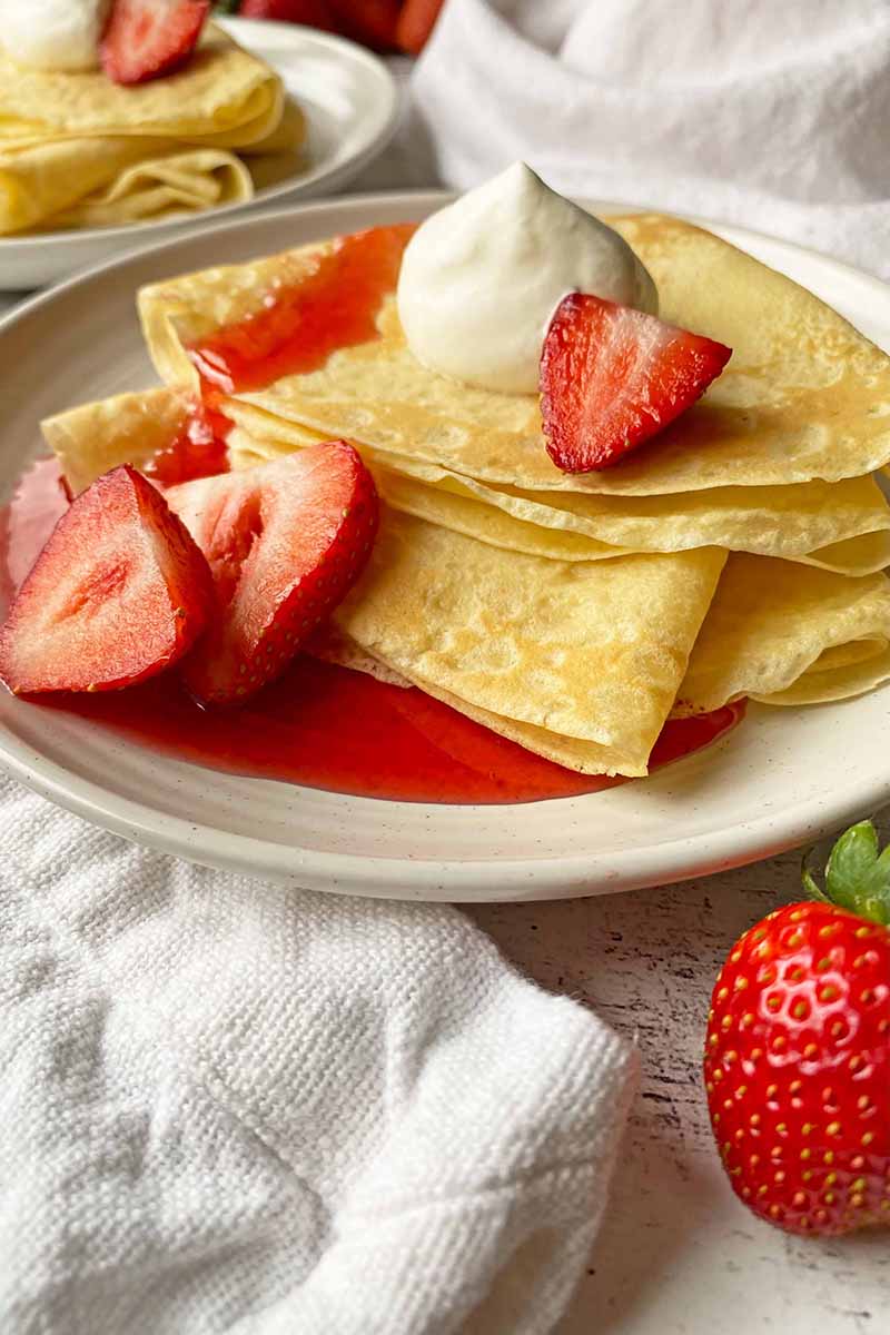 Vertical image of a stack of folded French-style thin pancakes on white plates topped with a dollop of whipped cream and sliced fruit next to a white napkin.
