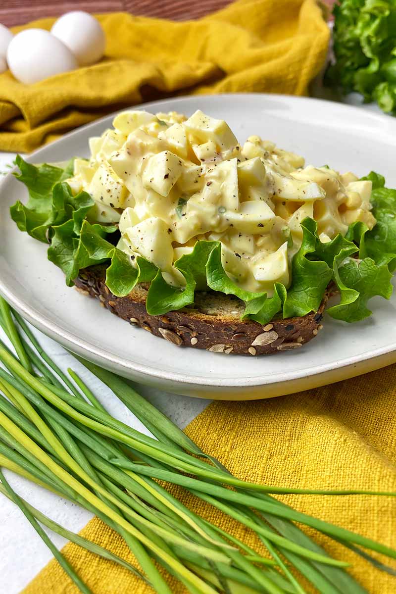 Vertical image of an open-faced sandwich topped with lettuce and a creamy mix of hard-boiled yolks and whites on a white plate next to chives and a yellow towel.