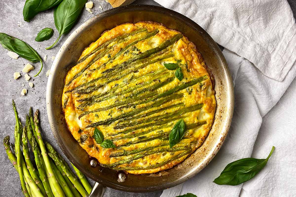 Horizontal top-down image of a whole frittata in a pan topped with asparagus spears and basil leaves.
