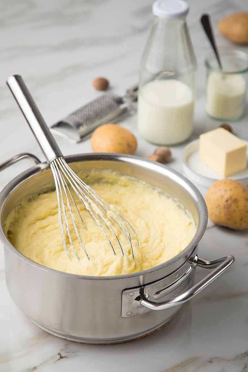 Vertical image of preparing mashed potatoes in a pot surrounded by ingredients on a marble work surface.