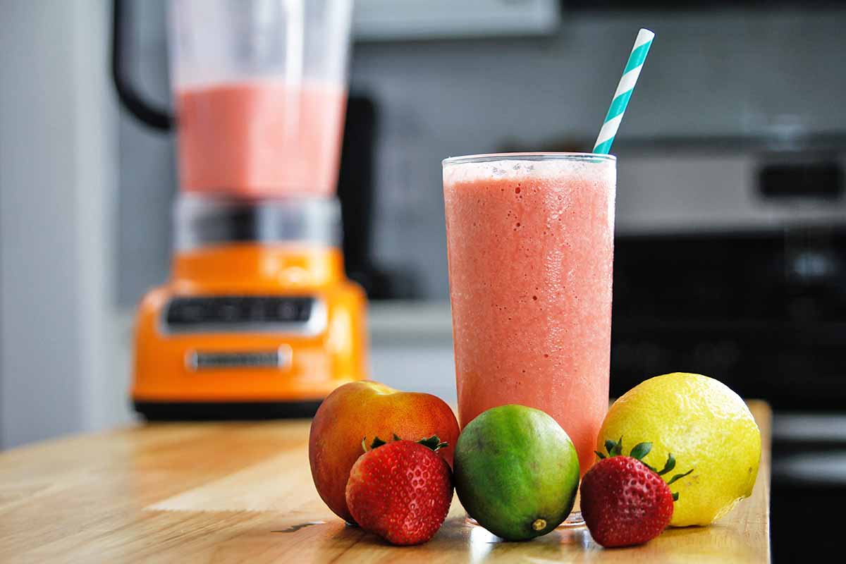 Horizontal image of a bright red shake in a glass cup with a striped straw next to fresh citrus and berries on a counter.