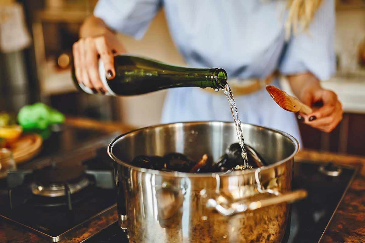 Horizontal image of pouring liquid from a bottle into a pot with mussels on the stovetop.