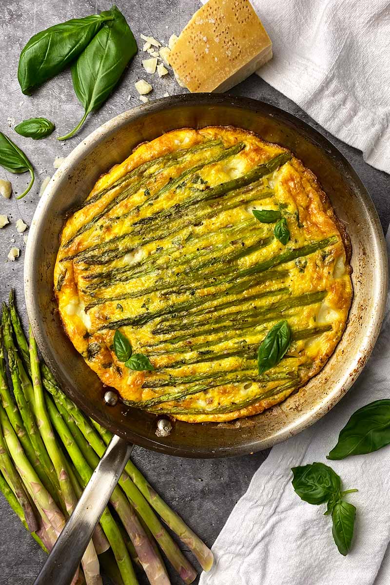 Vertical top-down image of a whole frittata in a skillet with asparagus spears and basil leaves.