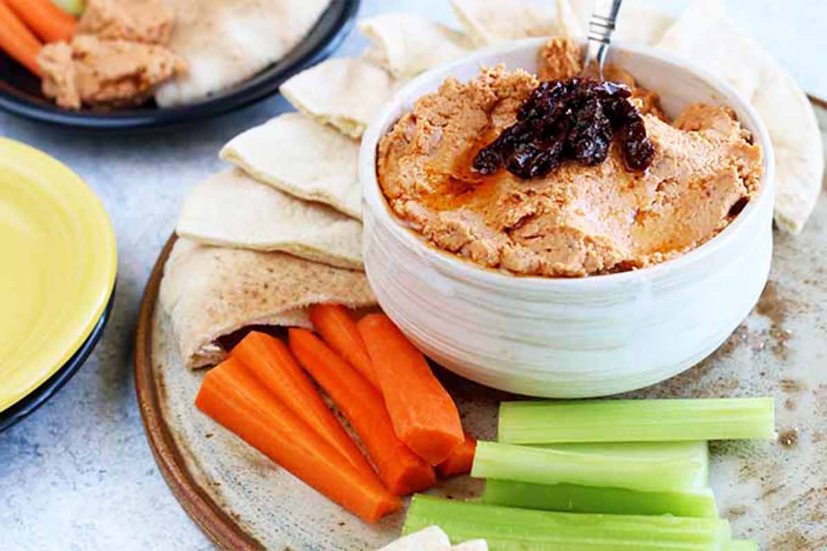 Horizontal image of a sun-dried tomato bean puree in a bowl on a large plate surrounded by celery, carrot, and pita.