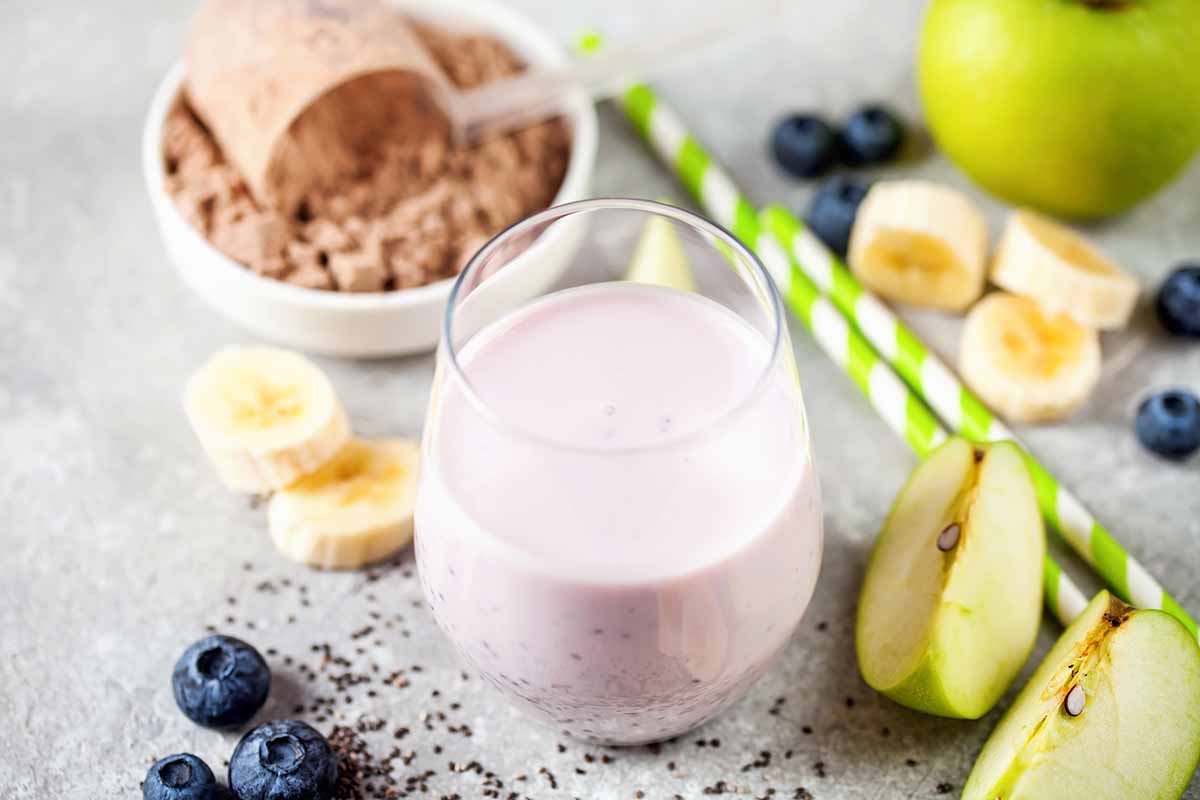 Horizontal image of a berry shake in a glass cup next to protein powder, straws, and assorted fresh fruit.