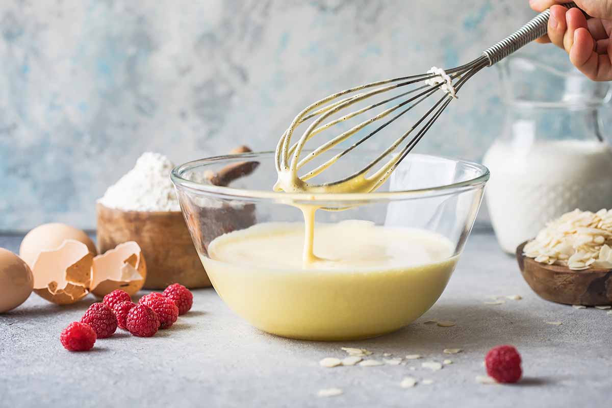 Horizontal image of making a thin batter in a glass bowl on a light gray surface next to raspberries, milk, almonds, flour, and eggs.