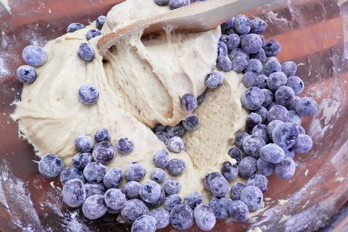 Horizontal image of folding in lightly dusted blueberries into a batter with a wooden spoon.