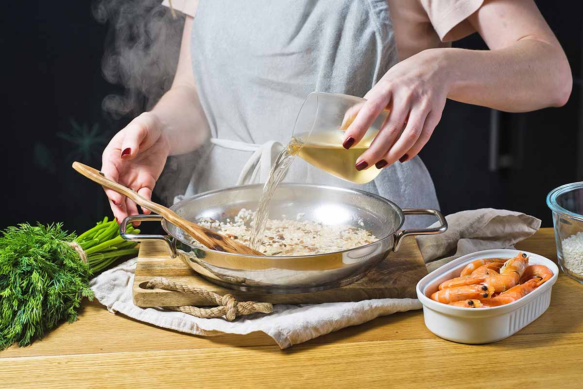 Horizontal image of a chef pouring a liquid into risotto in a pan, next to fresh herbs and shrimp.