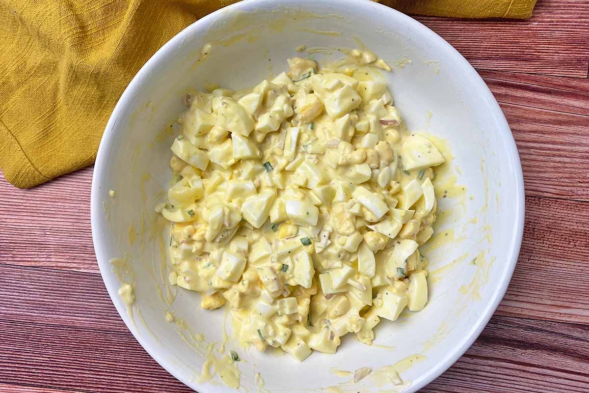 Horizontal image of a creamy, mayo-based mix with roughly chopped yolks and whites in a large white bowl.