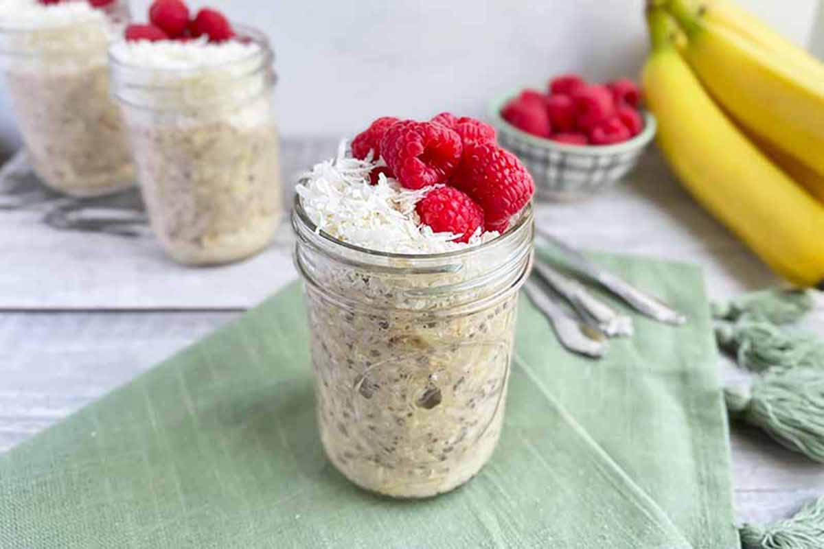 Horizontal image of overnight oats in glass jars topped with coconut and raspberries on a blue towel with bananas in the background.