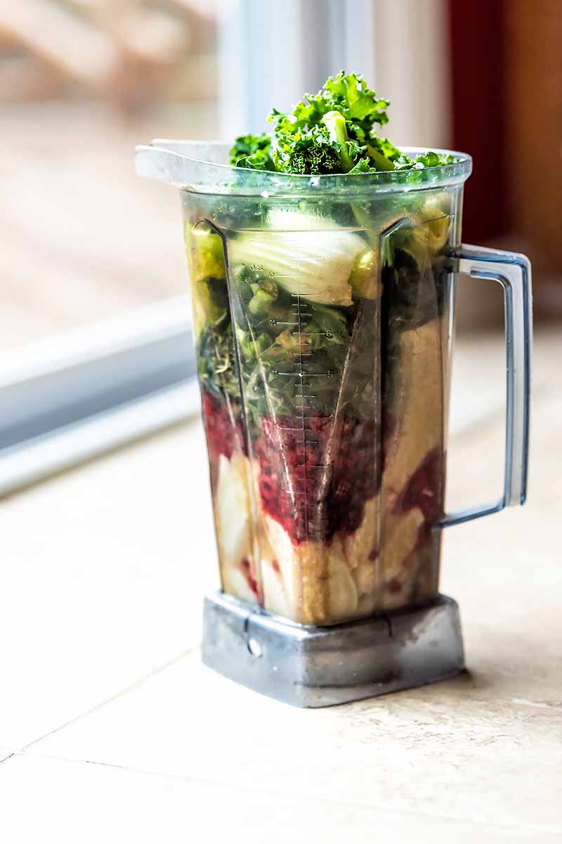 Vertical image of a plastic pitcher overfilled with assorted fruits and vegetables on a counter next to a window.