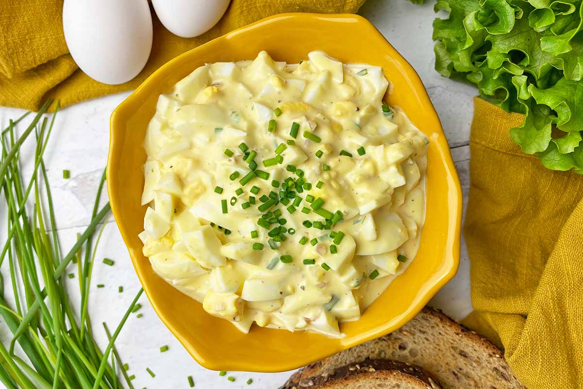 Horizontal top-down image of a yellow bowl filled with a creamy dish with chopped whites and yolks topped with chives next to bread slices and a yellow towel.