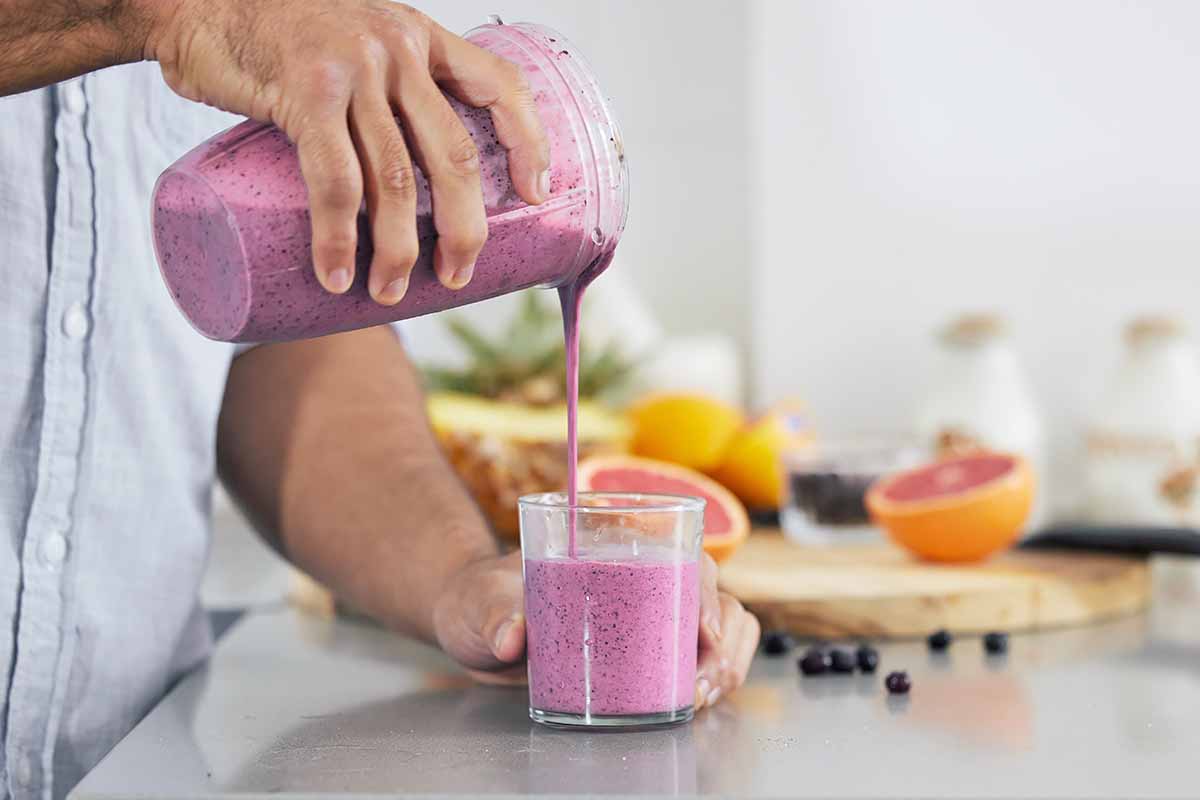 Horizontal image of a man pouring a berry shake into a glass on a countertop in front of fresh fruit.