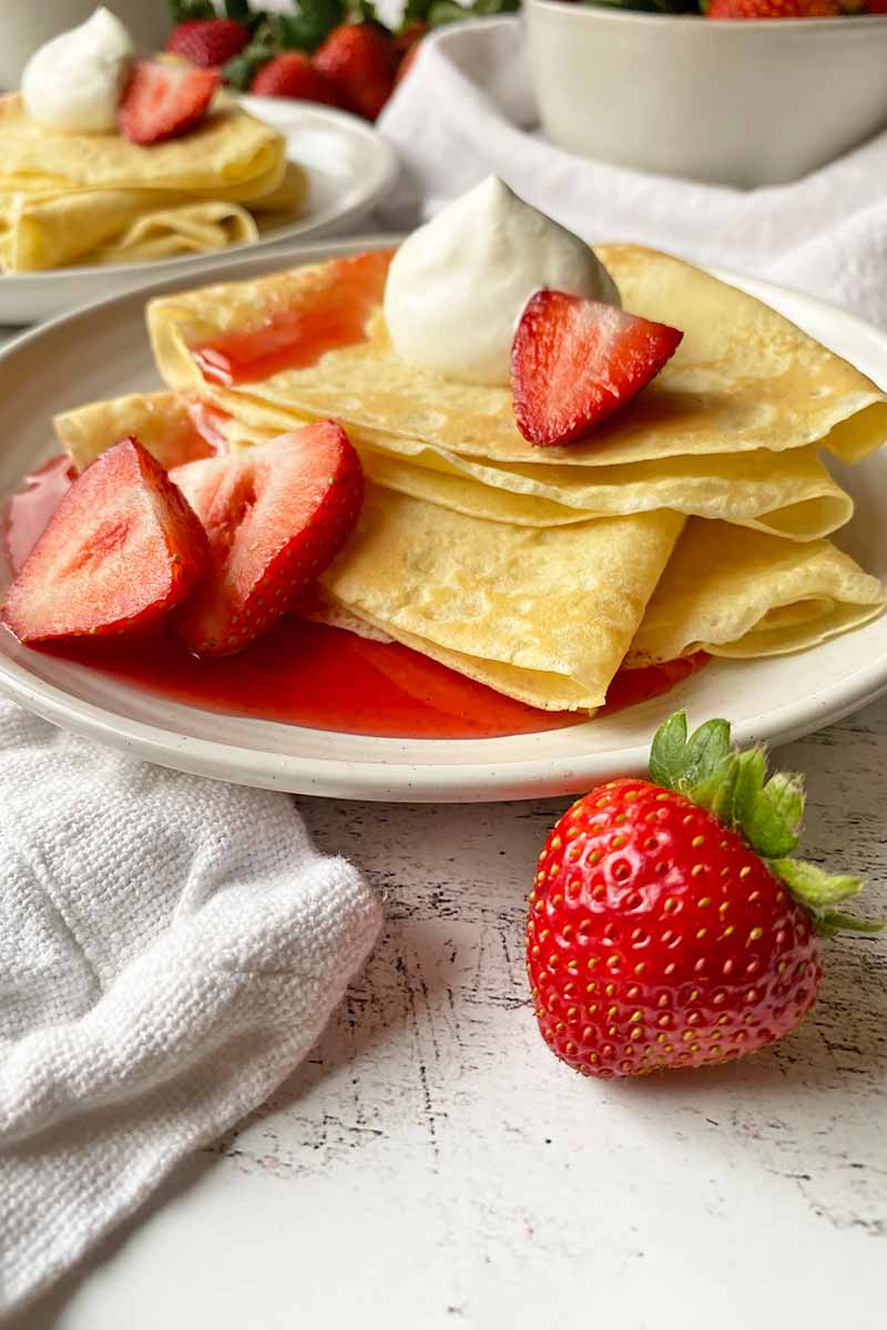 Vertical image of two white plates with a stack of folded thin pancakes topped with a dollop of whipped cream, sauce, and fresh fruit.