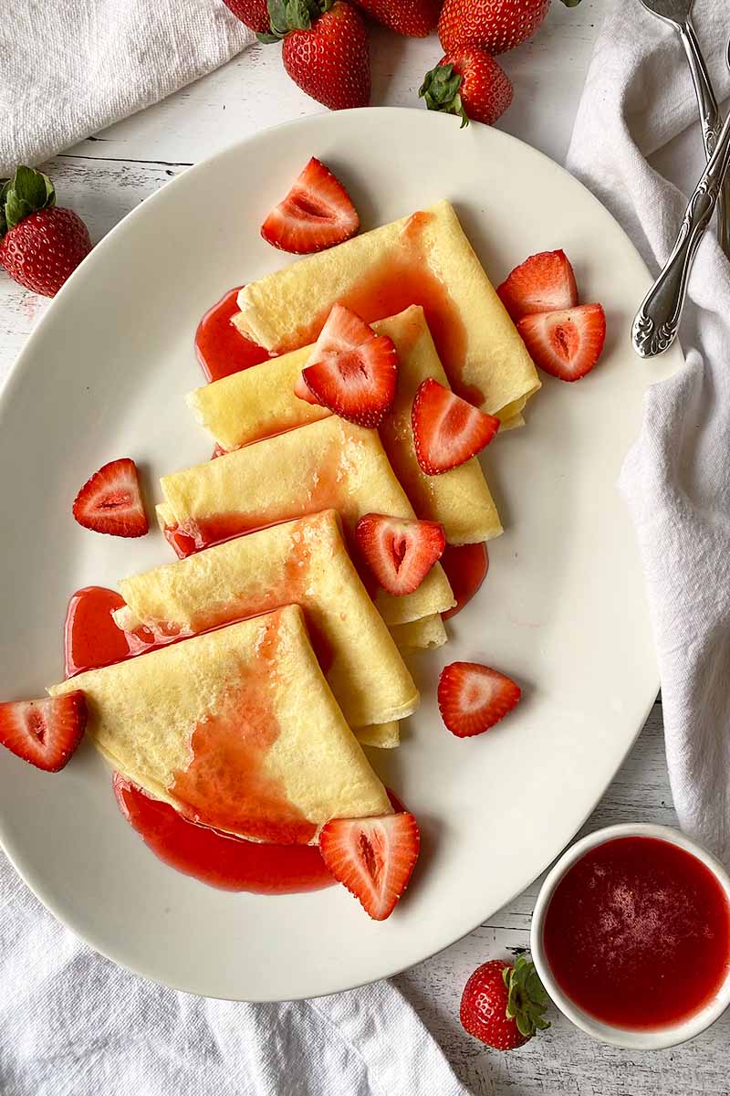 Vertical top-down image of shingled thin pancakes on a platter, topped with fresh fruity and sryup.