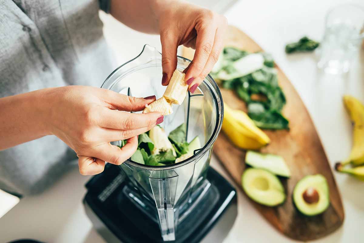 Horizontal image of breaking a fresh banana in half to put in a pitcher, next to a counter with sliced avocados and greens.