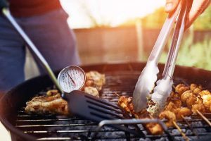 Horizontal image of a thermometer inserted in meat while preparing a BBQ outside while using tongs.