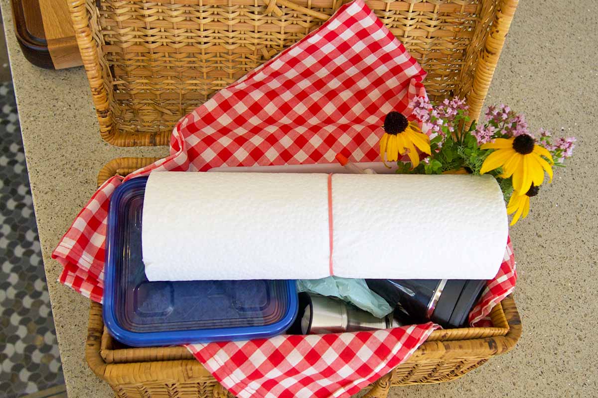 Horizontal image of a perfectly packed basket with paper towels, airtight containers of food, flowers, a towel, and bottled drinks.