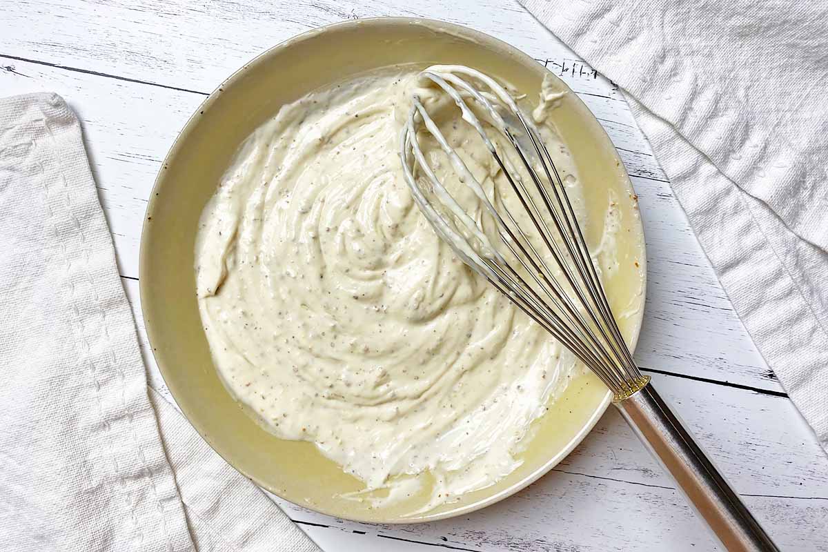 Horizontal image of whisking together a creamy white dressing in a shallow bowl.