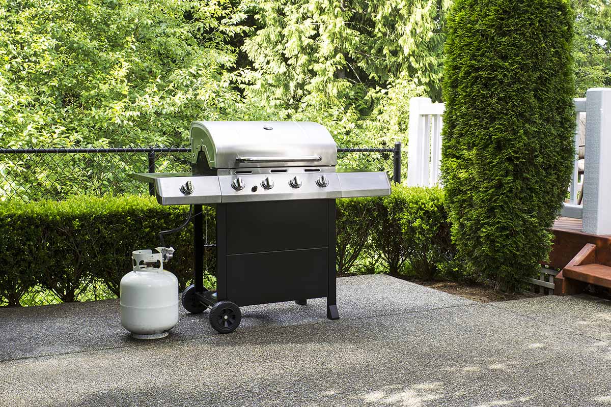 Horizontal image of a large propane gas grill on a patio outside, with shrubbery in the background.