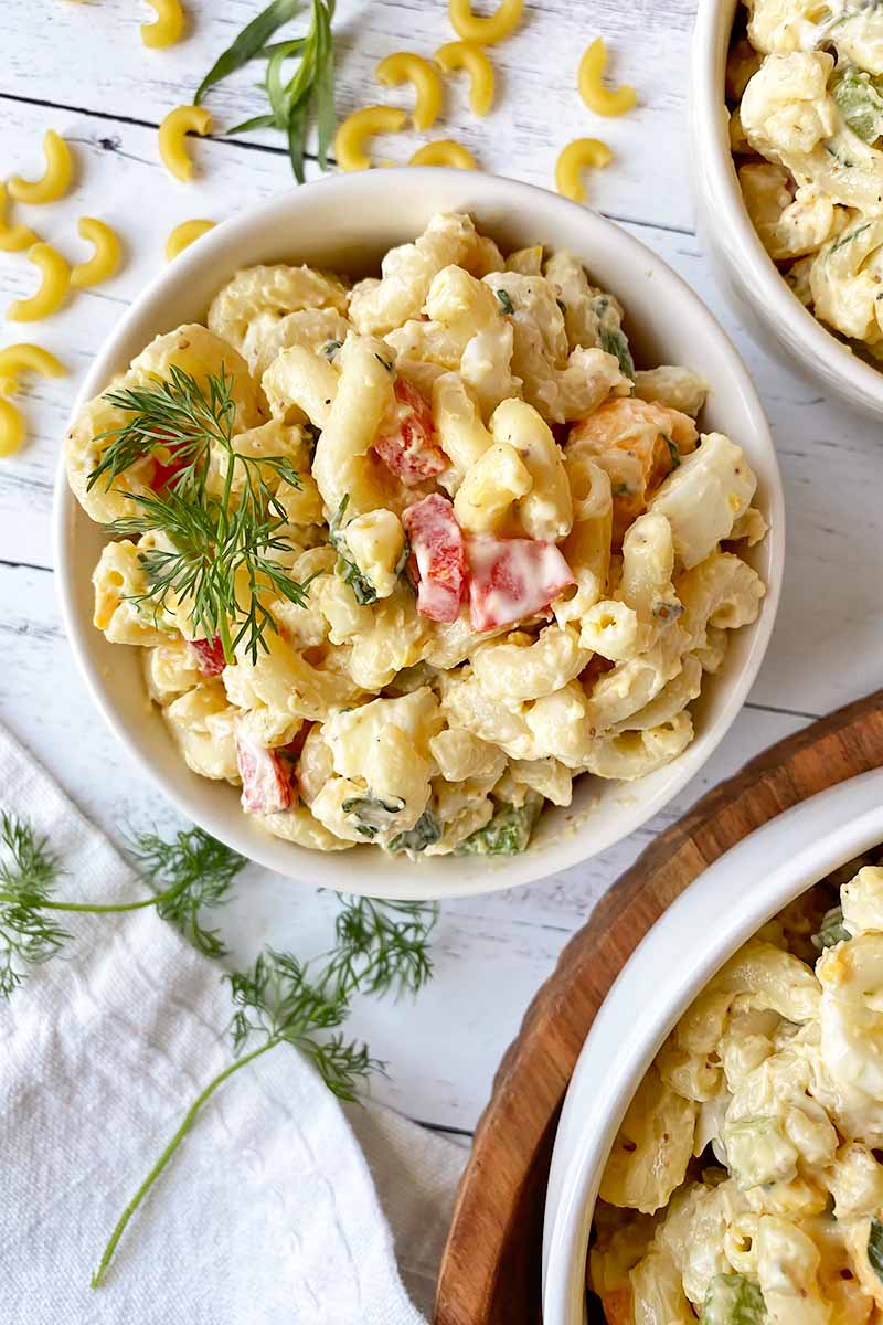 Vertical top-down image of assorted white bowls filled with a chilled macaroni recipe with assorted vegetables, garnished with dill fronds.