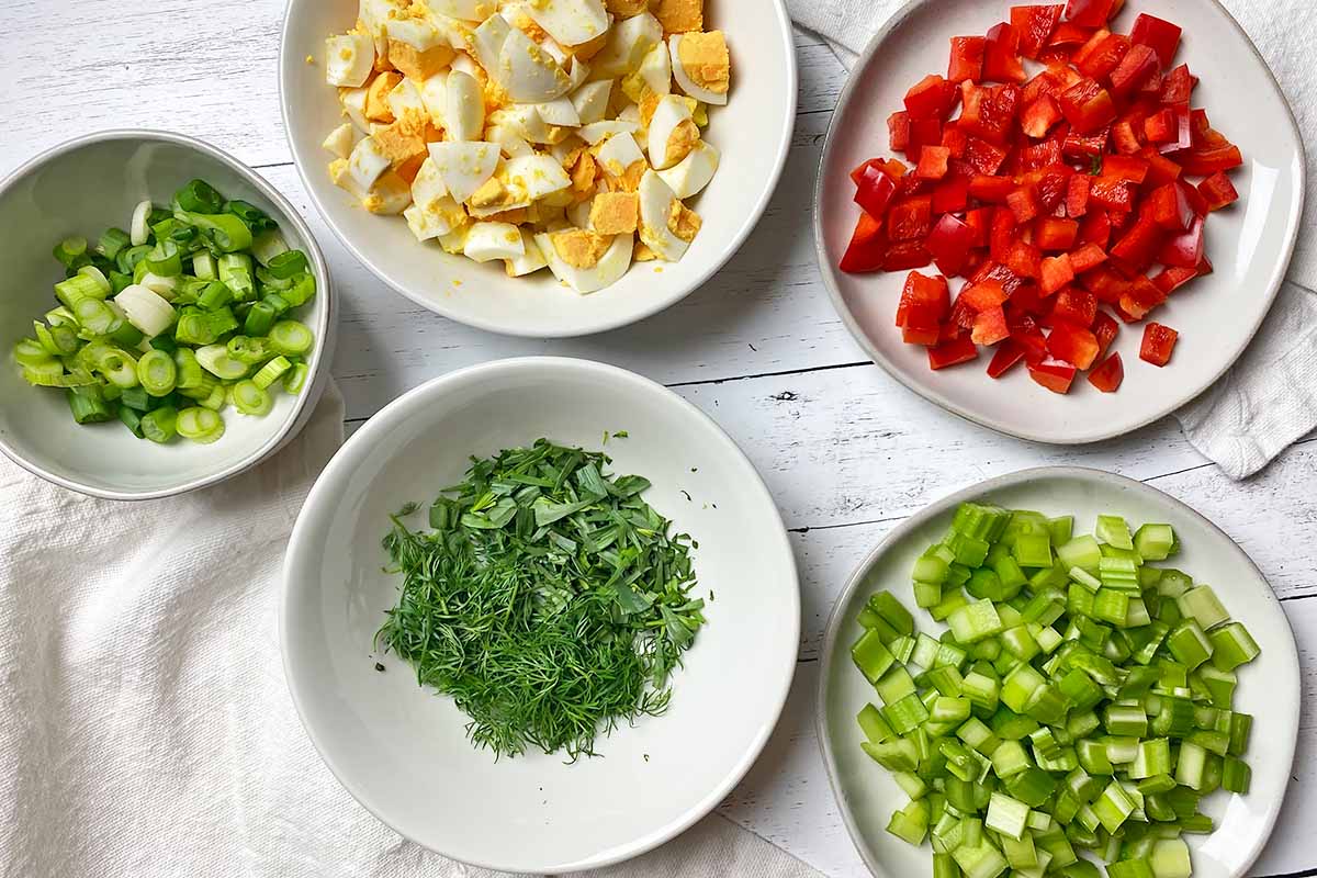 Horizontal image of assorted chopped vegetables and hard-boiled eggs on white plates.