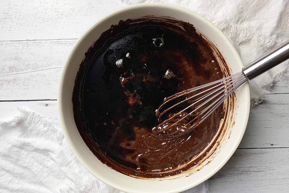 Horizontal image of mixing together a dark, thick mixture with a whisk in a large white bowl.