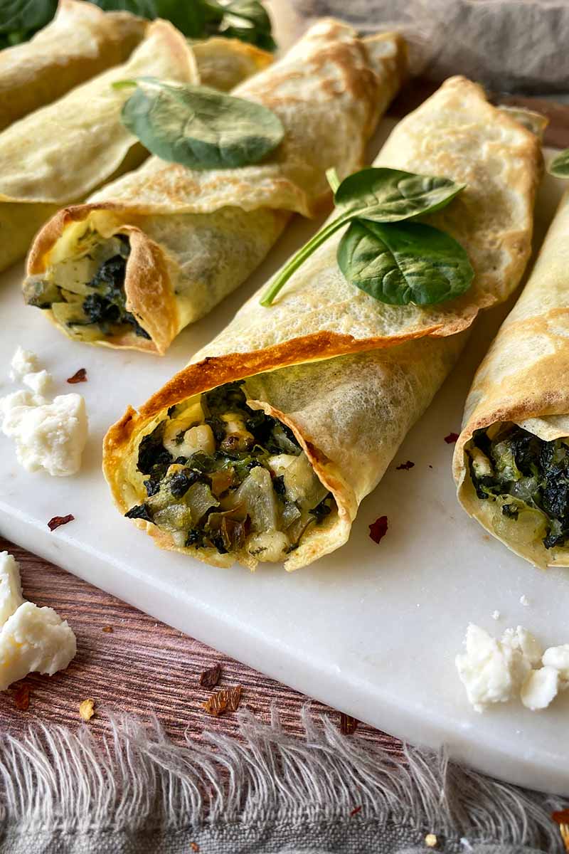 Vertical image of rolled thin pancakes with crispy edges and a greens, onion, and cheese stuffing on a serving platter.