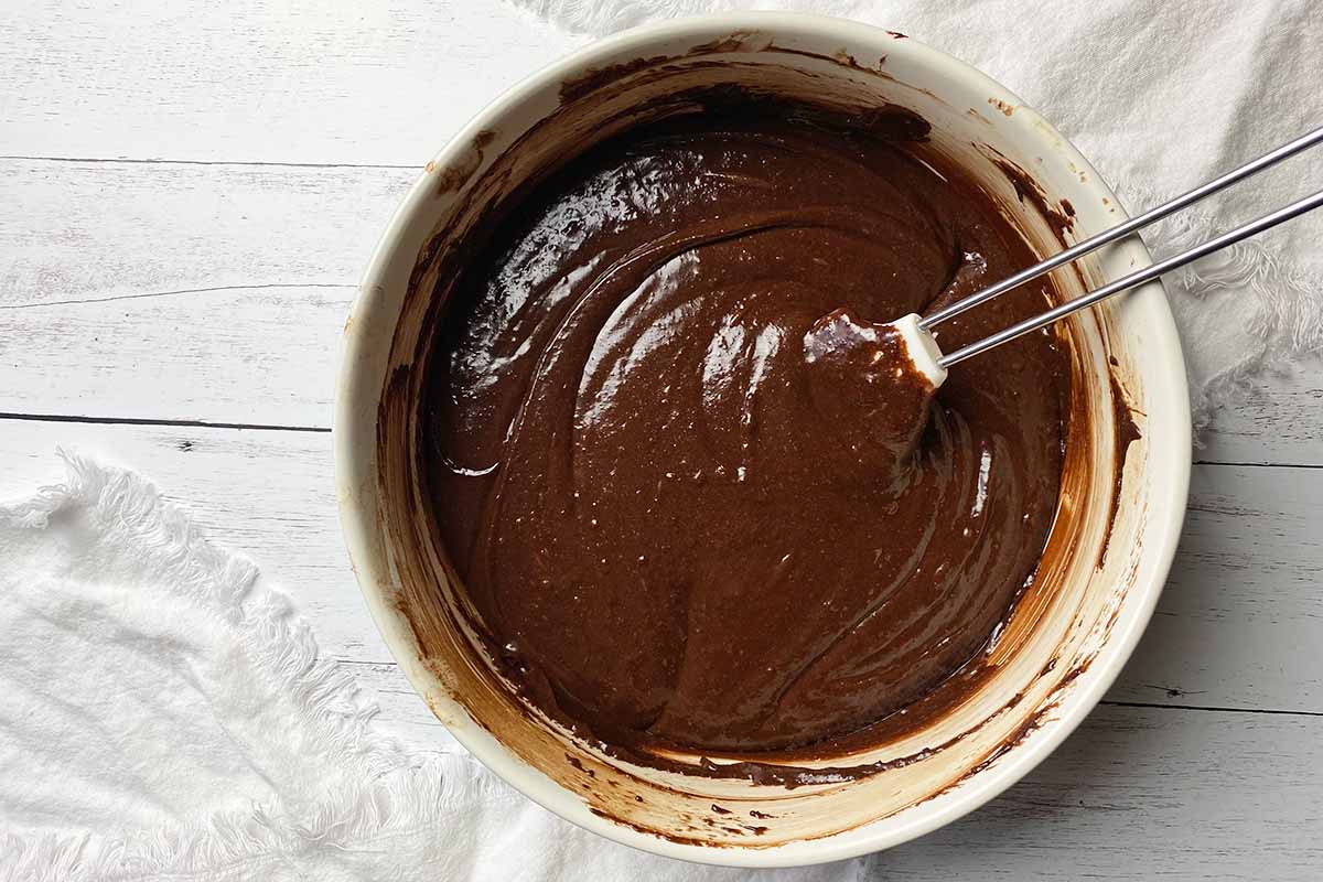 Horizontal image of mixing together a thick, deep brown mixture in a large white bowl with a spatula.