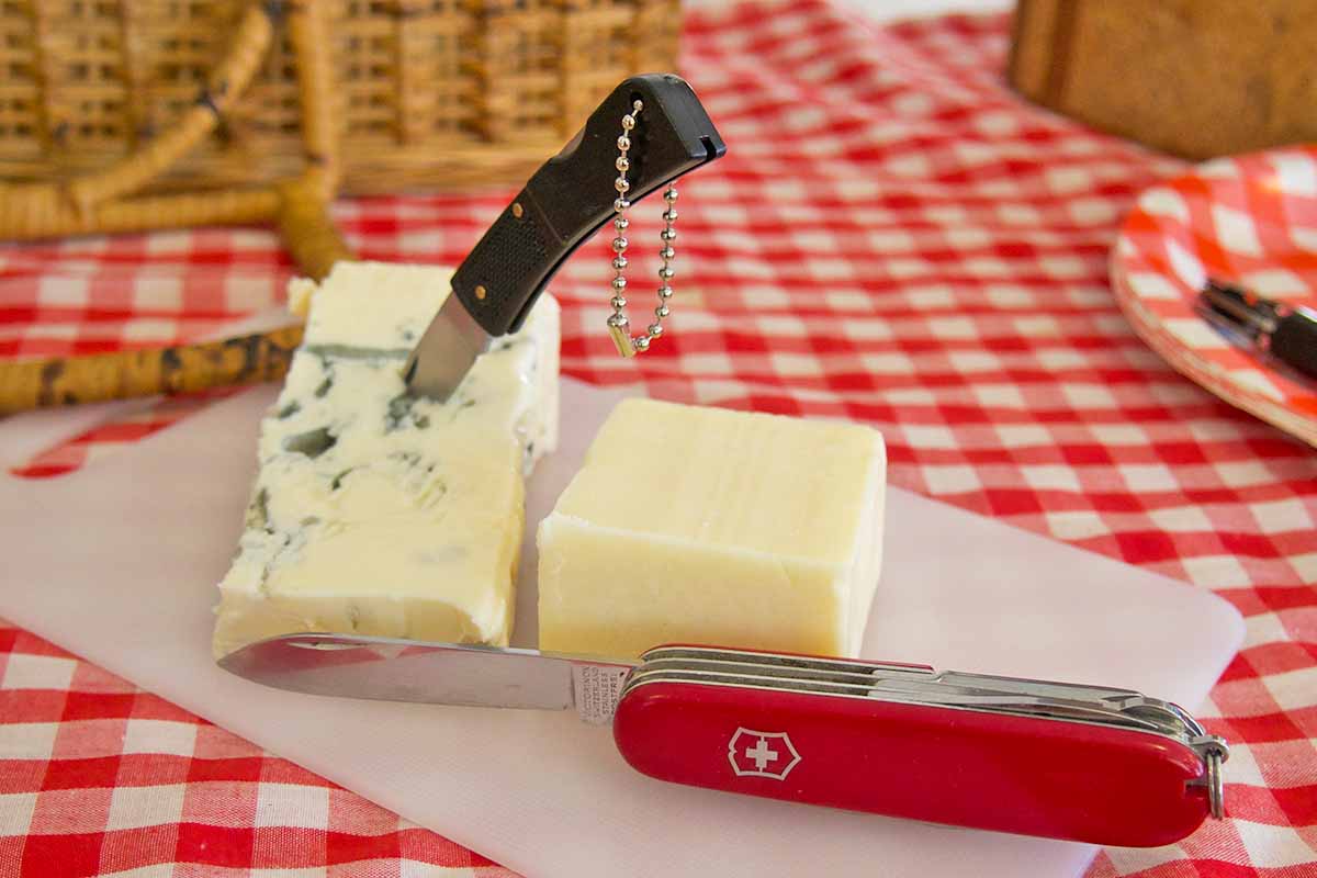 Horizontal image of using travel knives to cut cheese on a checkered table mat outside.